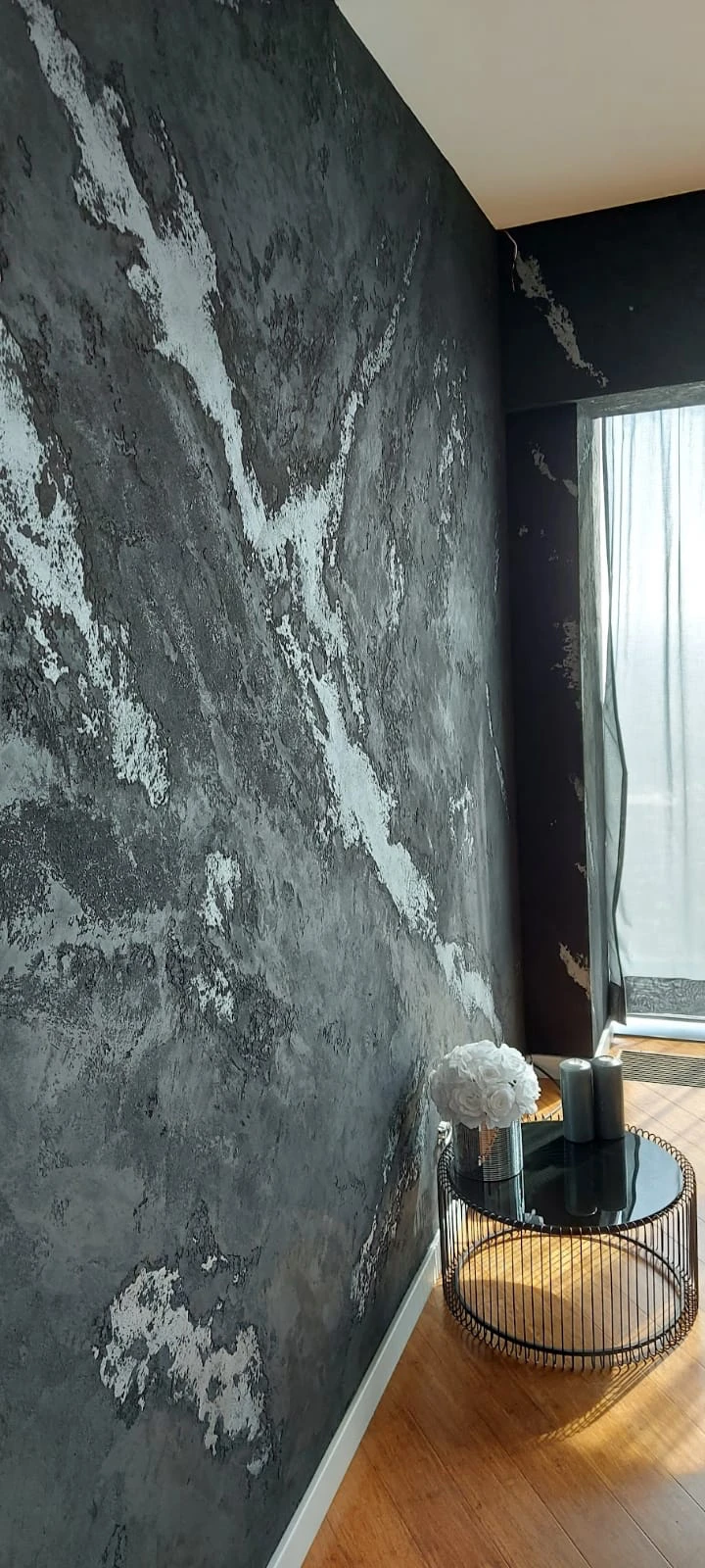 Ściana z Tynkami strukturalnymi w grafitowym odcieniu z białymi przetarciami obok stolika ze szklanym blatem i białymi kwiatami