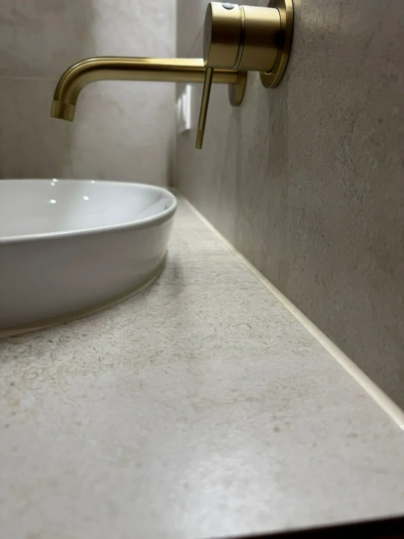 Złota ścienna bateria nad białą ceramiczną umywalką na kamiennym blacie, Umywalki