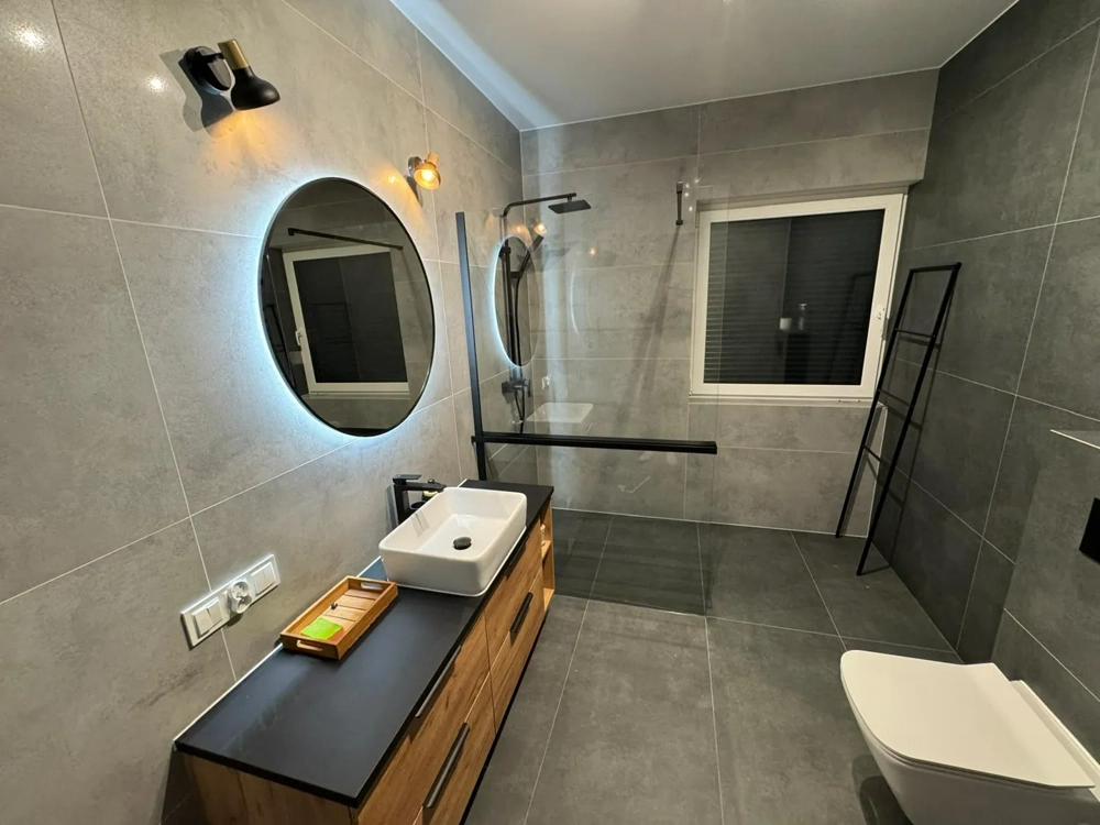 Łazienka po remoncie z dużymi szarymi płytkami, szklaną kabiną, drewnianą szafką i umywalką Remont łazienki, Układanie płytek