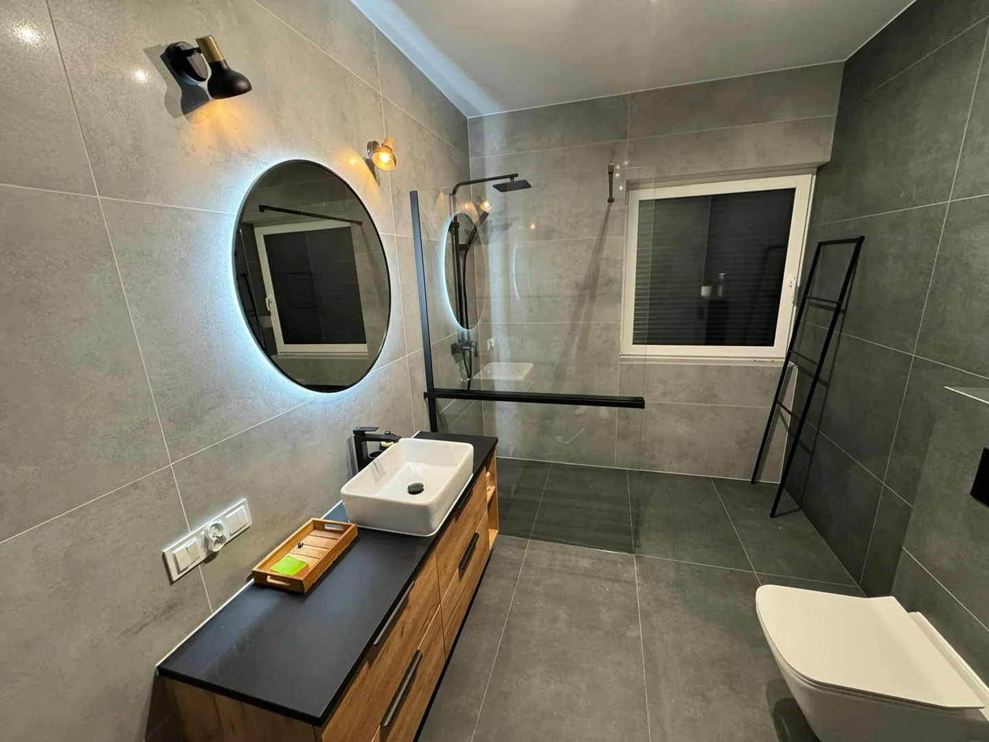 Nowoczesny remont łazienki z dużymi szarymi płytkami, podwieszanym sedesem, drewnianą szafką pod umywalką i oświetlonym lustrem