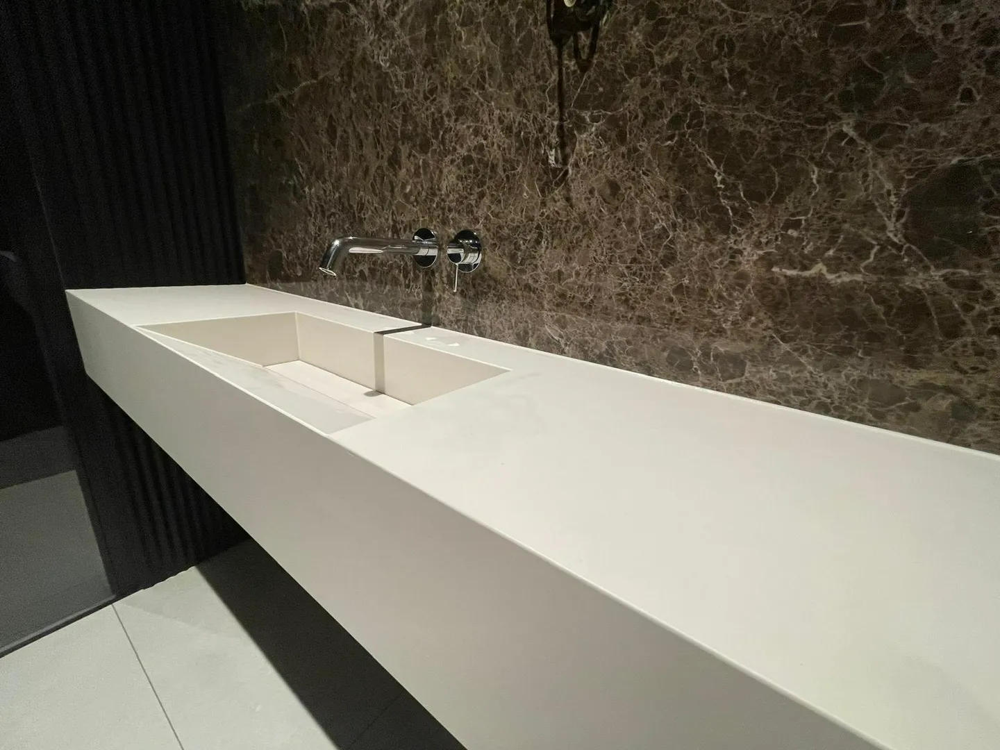 Biała minimalistyczna umywalka wkomponowana w blat z chromowaną baterią na tle brązowej marmurowej ściany – Umywalki