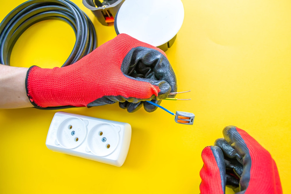 Ręce w czerwonych rękawicach montują przewody instalacji elektrycznej przy gniazdku na żółtym tle